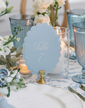 A styled wedding table setting featuring a light blue scalloped table number sign reading 'Table 7' on a gold stand, surrounded by white and pale blue floral arrangements, textured blue glassware, white plates, and a blue napkin.