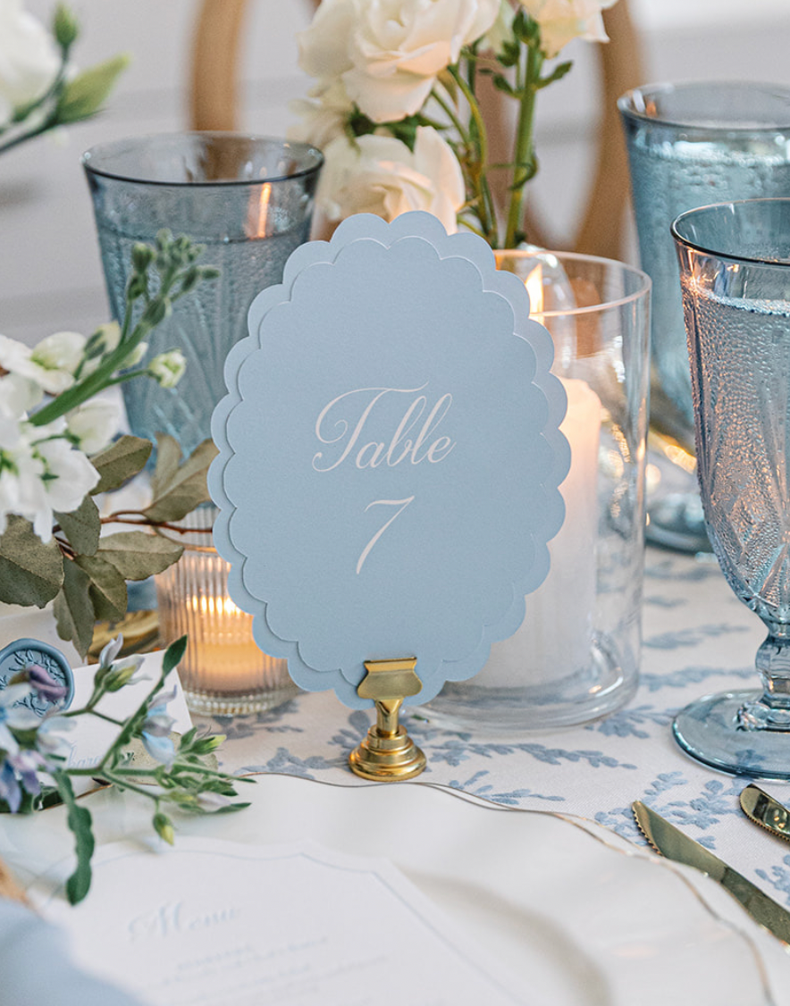 A styled wedding table setting featuring a light blue scalloped table number sign reading 'Table 7' on a gold stand, surrounded by white and pale blue floral arrangements, textured blue glassware, white plates, and a blue napkin.